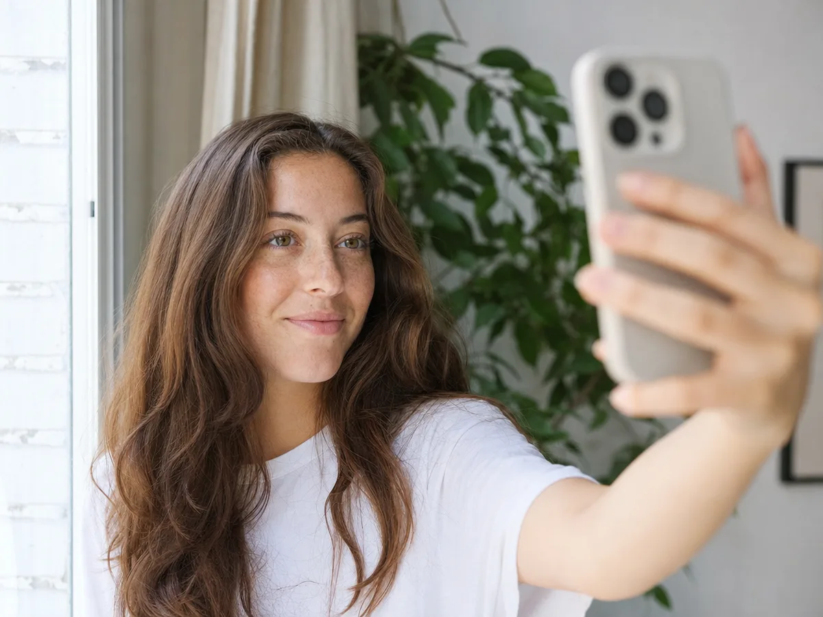 A woman with long chestnut-brown hair takes a selfie facing a bright window at home
